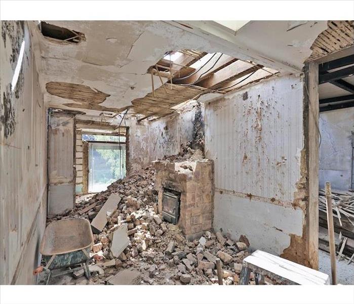 A long narrow room in an older home with a fireplace after a fire; bricks from a crumbled chimney scattered; large window.